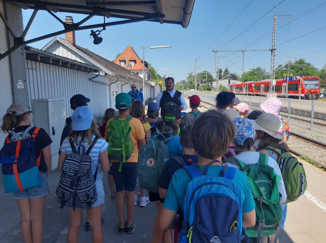 Hans-Jörg Gruppe von Kindern mit Rucksäcken wartet an einem Bahnhof auf eine Ansprache.
